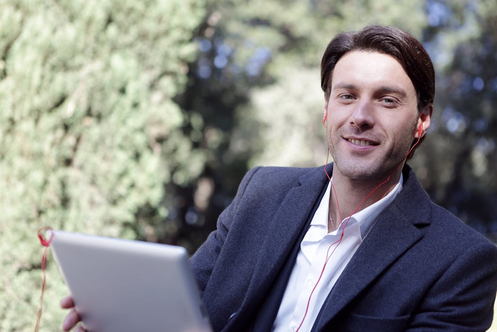 Smiling businessman with headphones using a tablet in a sunny outdoor setting.