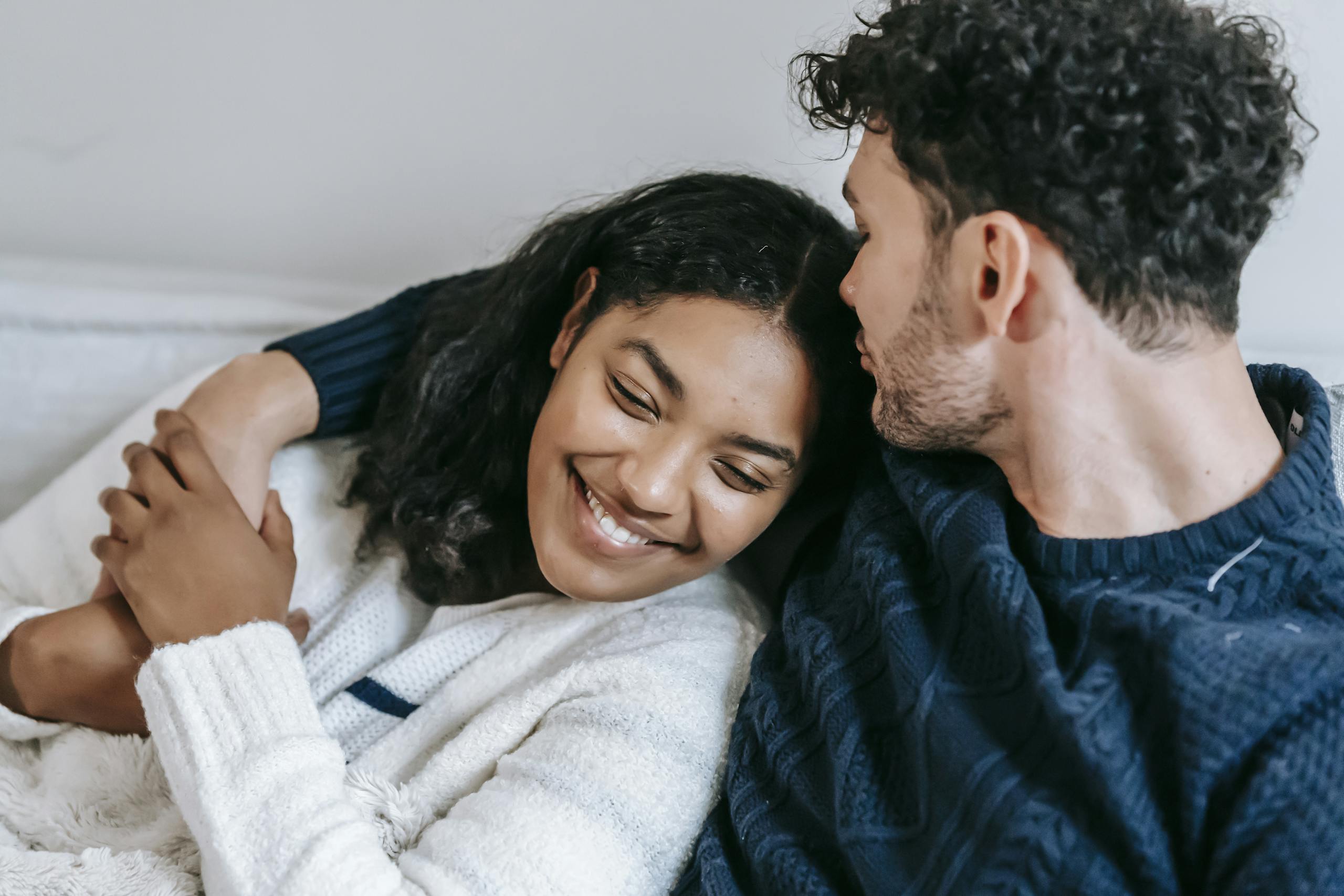 Crop enamored young man kissing head of happy ethnic girlfriend while embracing each other and resting in sofa at home