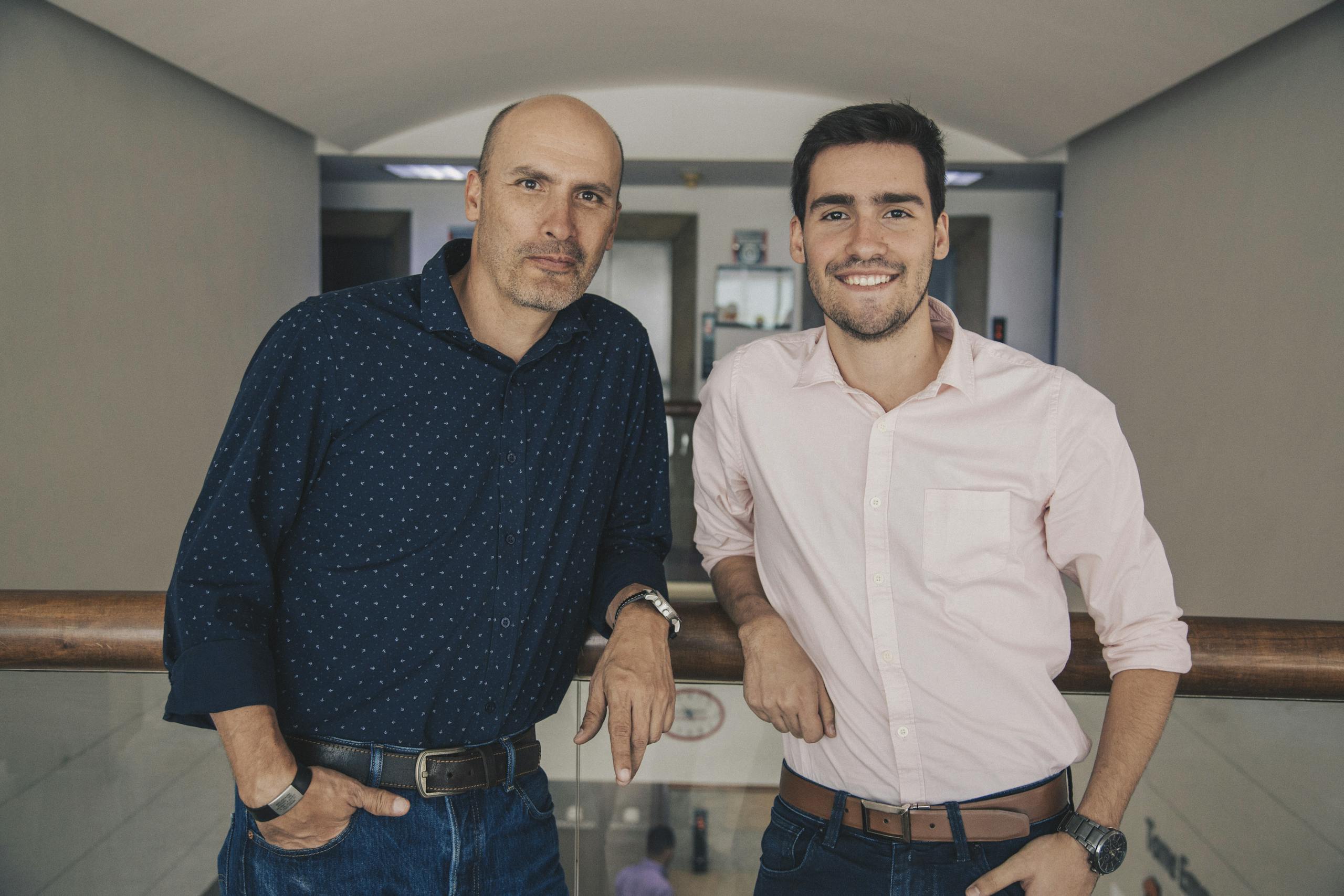 Smiling colleagues in shirts and jeans standing together and leaning on railing while looking at camera in white corridor